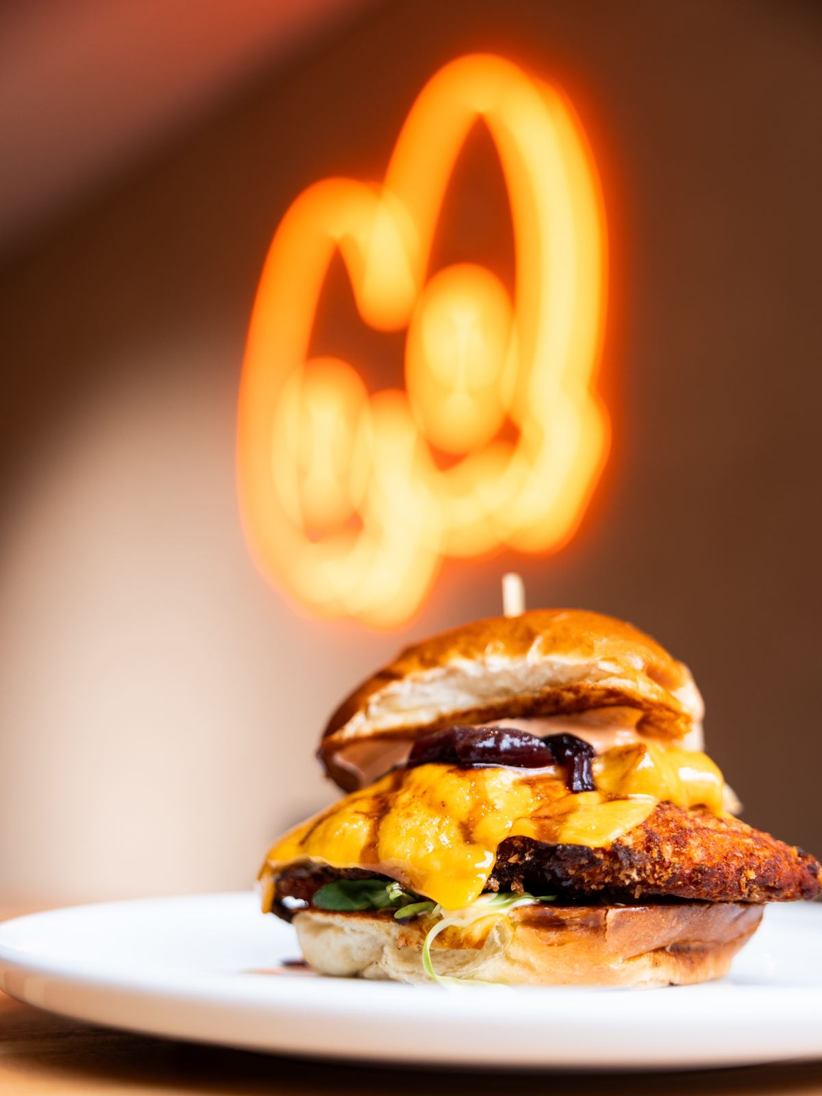 Crispy chicken burger s roztaveným čedarem, za ním svítí neon QUOQ